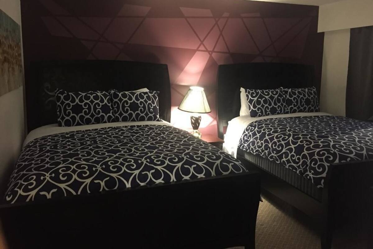 Quarto de hotel com duas camas, cada uma com colcha estampada, mesa de cabeceira com abajur e cabeceira com padrão geométrico.