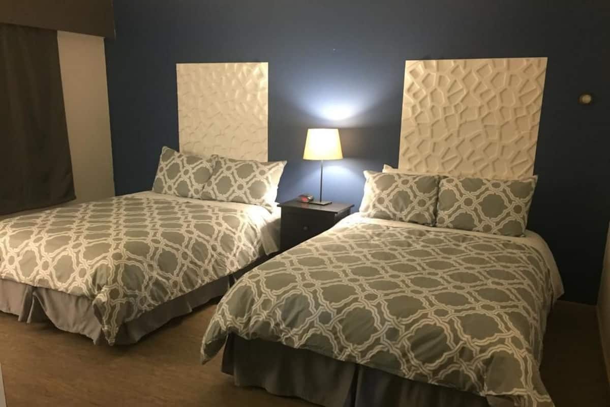 Duas camas com roupa de cama estampada, uma mesa de cabeceira com abajur e uma parede azul escura.