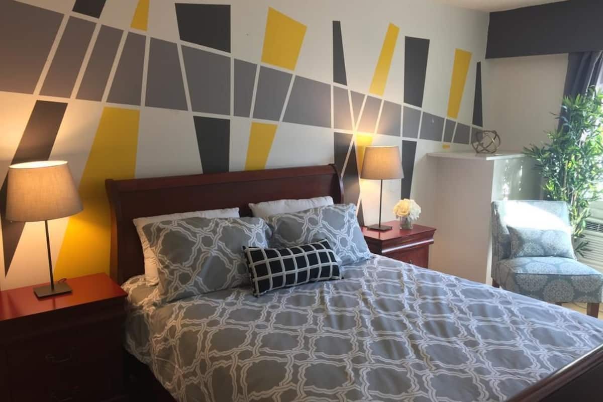 Cama bem arrumada com roupa de cama estampada, duas luminárias de cabeceira, um painel de cabeceira em madeira e uma parede com arte geométrica.