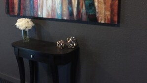 A room with a dark wall, a large abstract painting, a small table with a vase and two decorative items. - Hamilton Hotel (Williams Lake)