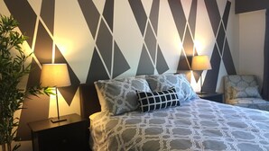 A modern hotel room with a geometric-patterned wall, a bed with patterned bedding, two bedside lamps, a chair, and a potted plant. - Hamilton Hotel (Williams Lake)