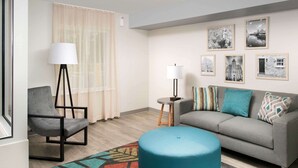 A modern living room with a grey sofa, a blue ottoman, a black chair, a floor lamp, and a patterned rug. - WoodSpring Suites Merrillville (Merrillville)