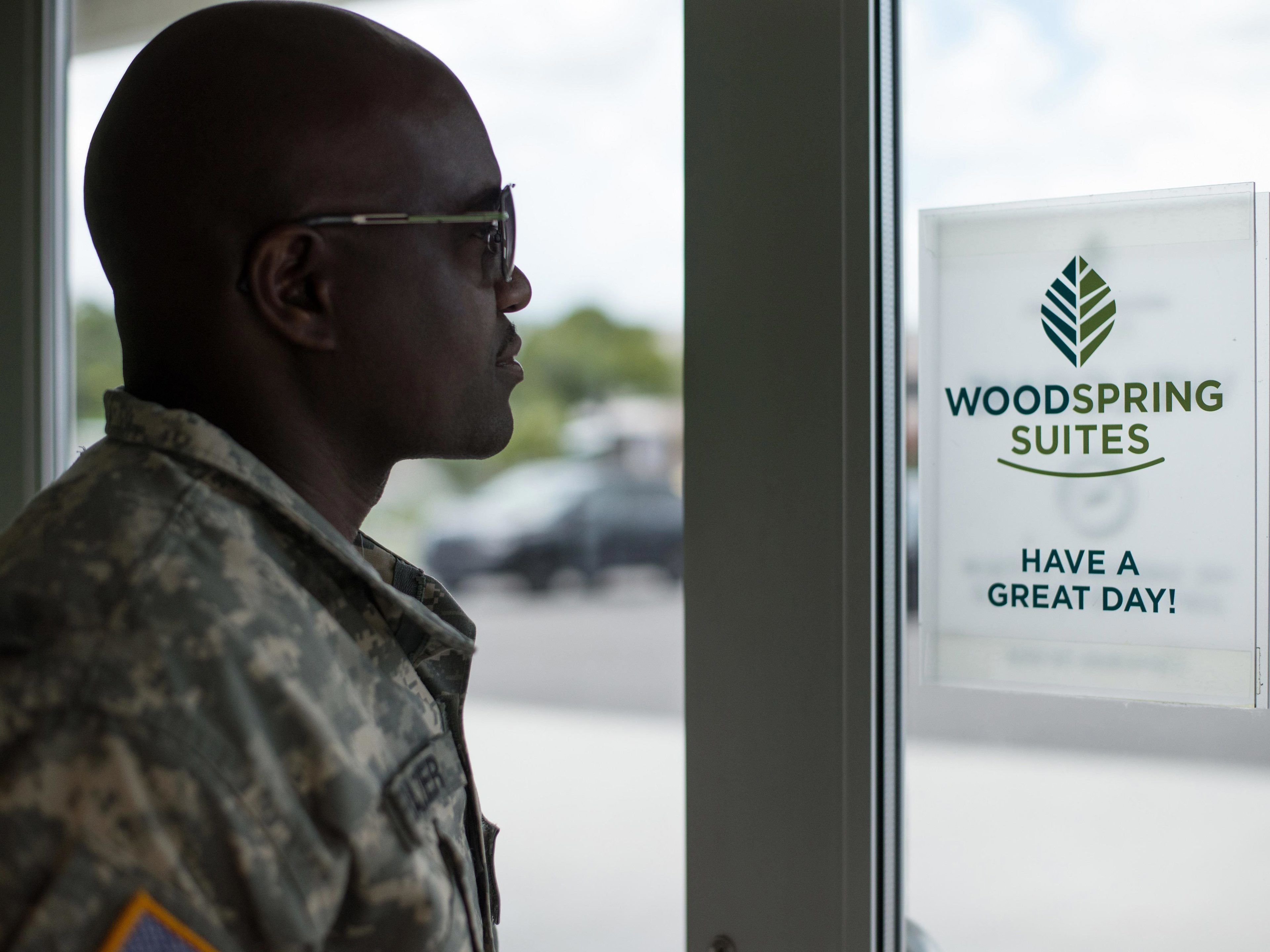 A person in military uniform standing in front of a window with a sign that reads "WOODSPRING SUITES HAVE A GREAT DAY!"