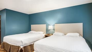 A room with two beds, each with a white bedspread and brown headboards. There is a nightstand with a lamp between the beds.