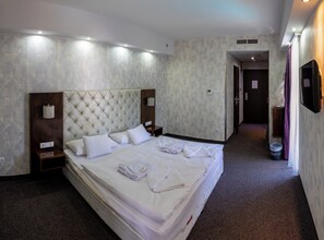 A hotel room with a large bed, a wooden nightstand, a wall-mounted TV, and a bathroom door in the background. - Bastya Wellness Hotel (Miskolc)