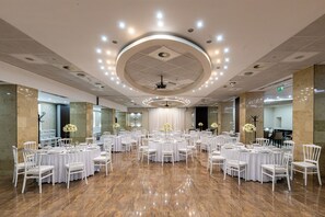 Un amplio salón de banquetes con varias mesas redondas y sillas blancas, un suelo de mármol pulido y un diseño de techo moderno con iluminación empotrada.
