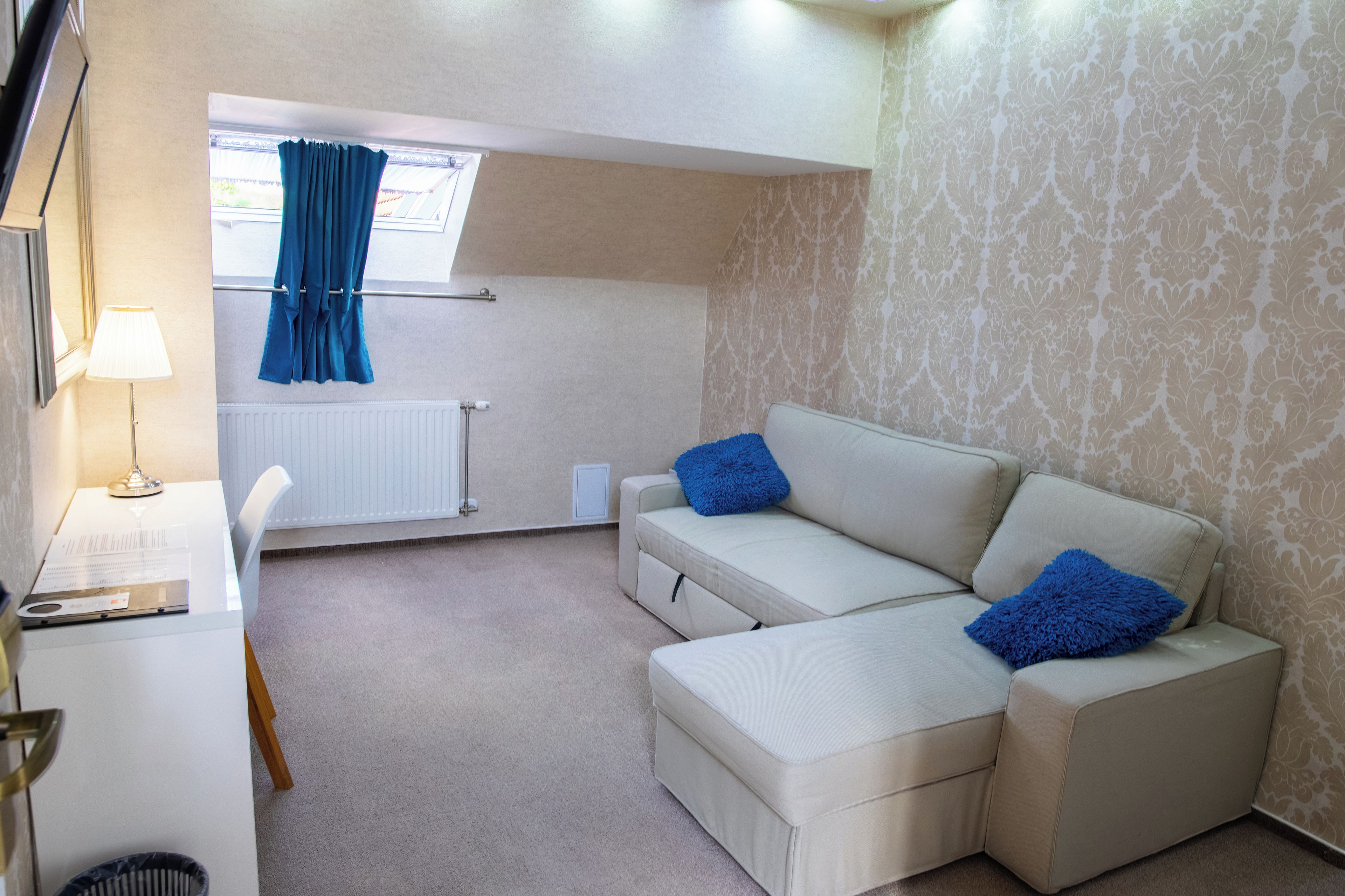A modern living room with a white sofa, two blue cushions, a white desk with a lamp, and a window with blue curtains.