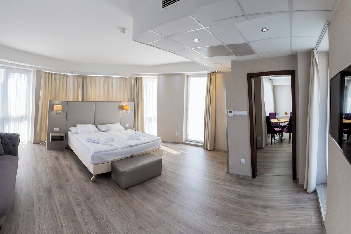 A modern hotel room with a curved ceiling, a large bed with a headboard, a sofa, and a dining area visible through an open door.