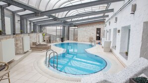 An indoor swimming pool with a curved design, surrounded by lounge chairs and a tiled floor. - Bastya Wellness Hotel (Miskolc)