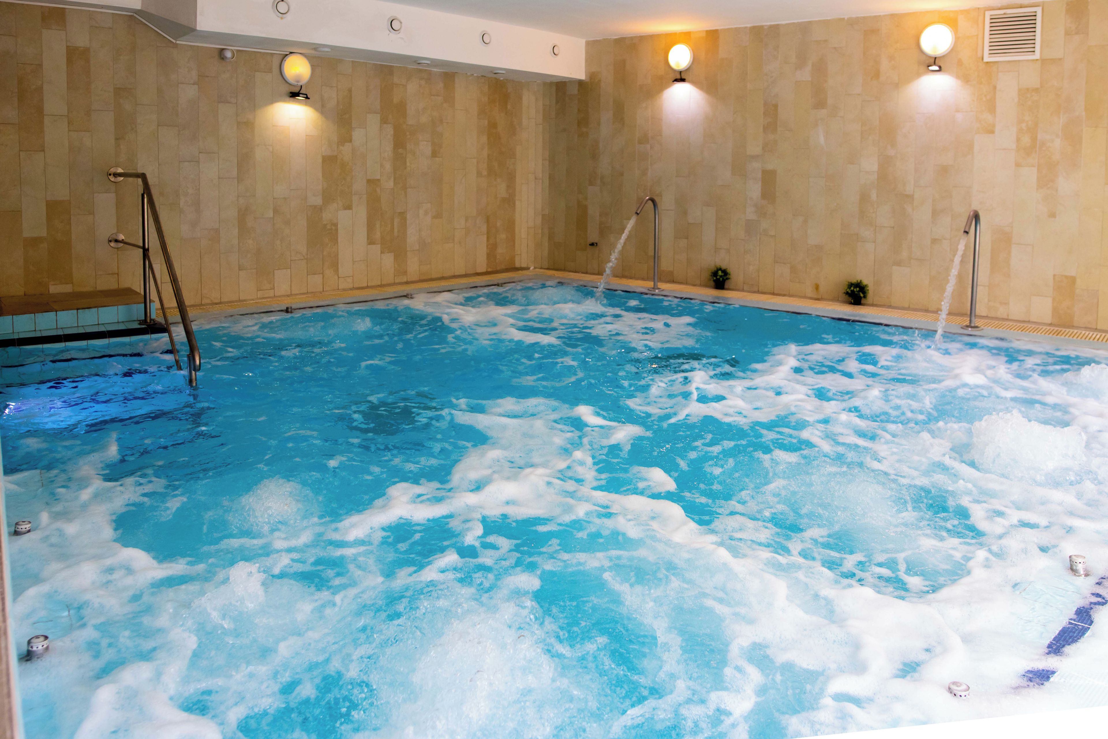 A large indoor swimming pool with clear water, a ladder on the left, and two wall-mounted lights.
