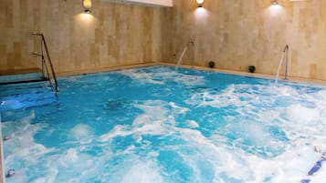 A large indoor swimming pool with clear water, a ladder on the left, and two wall-mounted lights.