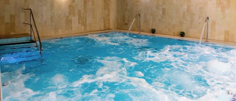 A large indoor swimming pool with clear water, a ladder on the left, and two wall-mounted lights.