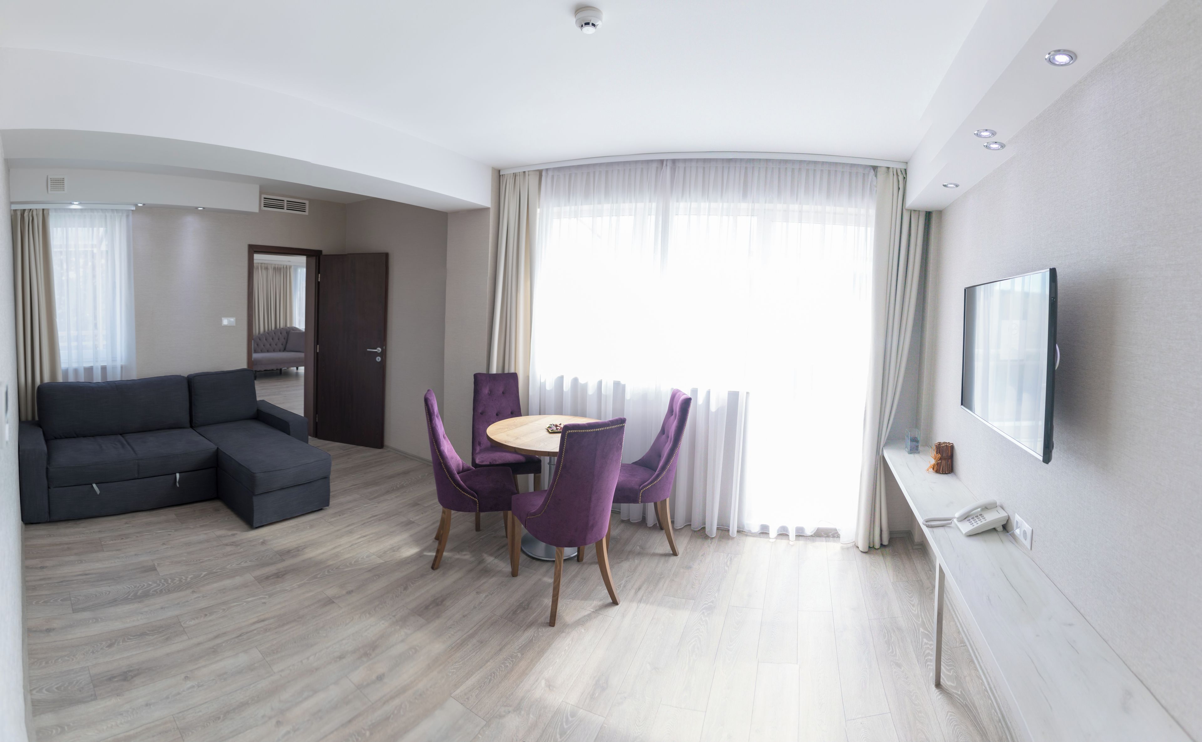A modern hotel room with a grey sectional sofa, a round dining table with purple chairs, a flat-screen TV mounted on the wall, and a large window with sheer curtains.