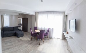 A modern hotel room with a grey sectional sofa, a round dining table with purple chairs, a flat-screen TV mounted on the wall, and a large window with sheer curtains. - Bastya Wellness Hotel (Miskolc)