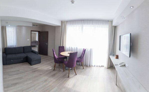 A modern hotel room with a grey sectional sofa, a round dining table with purple chairs, a flat-screen TV mounted on the wall, and a large window with sheer curtains. - Bastya Wellness Hotel (Miskolc)