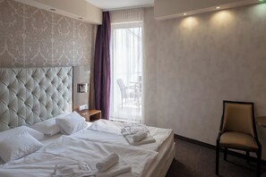A hotel room with a tufted headboard, a bed with white linens, a chair, and a view of the outdoors through a window with sheer curtains. - Bastya Wellness Hotel (Miskolc)