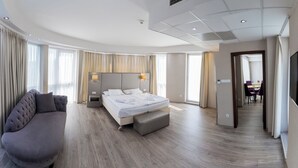 A modern hotel room with a curved ceiling, a large bed with a headboard, a sofa, and a dining area visible through an open door. - Bastya Wellness Hotel (Miskolc)