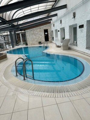 An indoor swimming pool with clear water, surrounded by tiled flooring and a high ceiling with skylights. - Bastya Wellness Hotel (Miskolc)