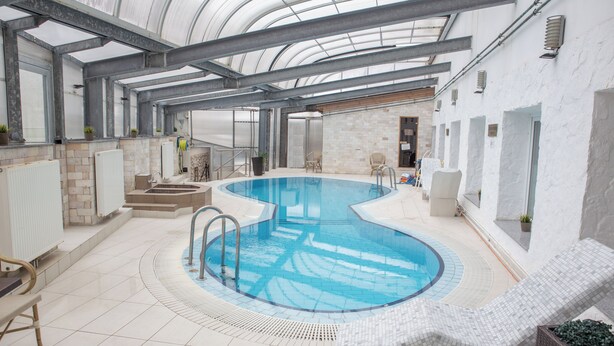 An indoor swimming pool with a curved design, surrounded by lounge chairs and a tiled floor. - Bastya Wellness Hotel (Miskolc)