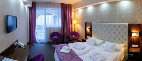 A hotel room with a large bed, two purple chairs, a desk with a phone, and a TV.