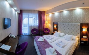 A hotel room with a large bed, two purple chairs, a desk with a phone, and a TV. - Bastya Wellness Hotel (Miskolc)