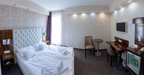 A hotel room with a large bed, a desk, a chair, a television, and a minibar. - Bastya Wellness Hotel (Miskolc)