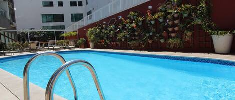 A swimming pool with metal railings, surrounded by a building with a red wall and potted plants.