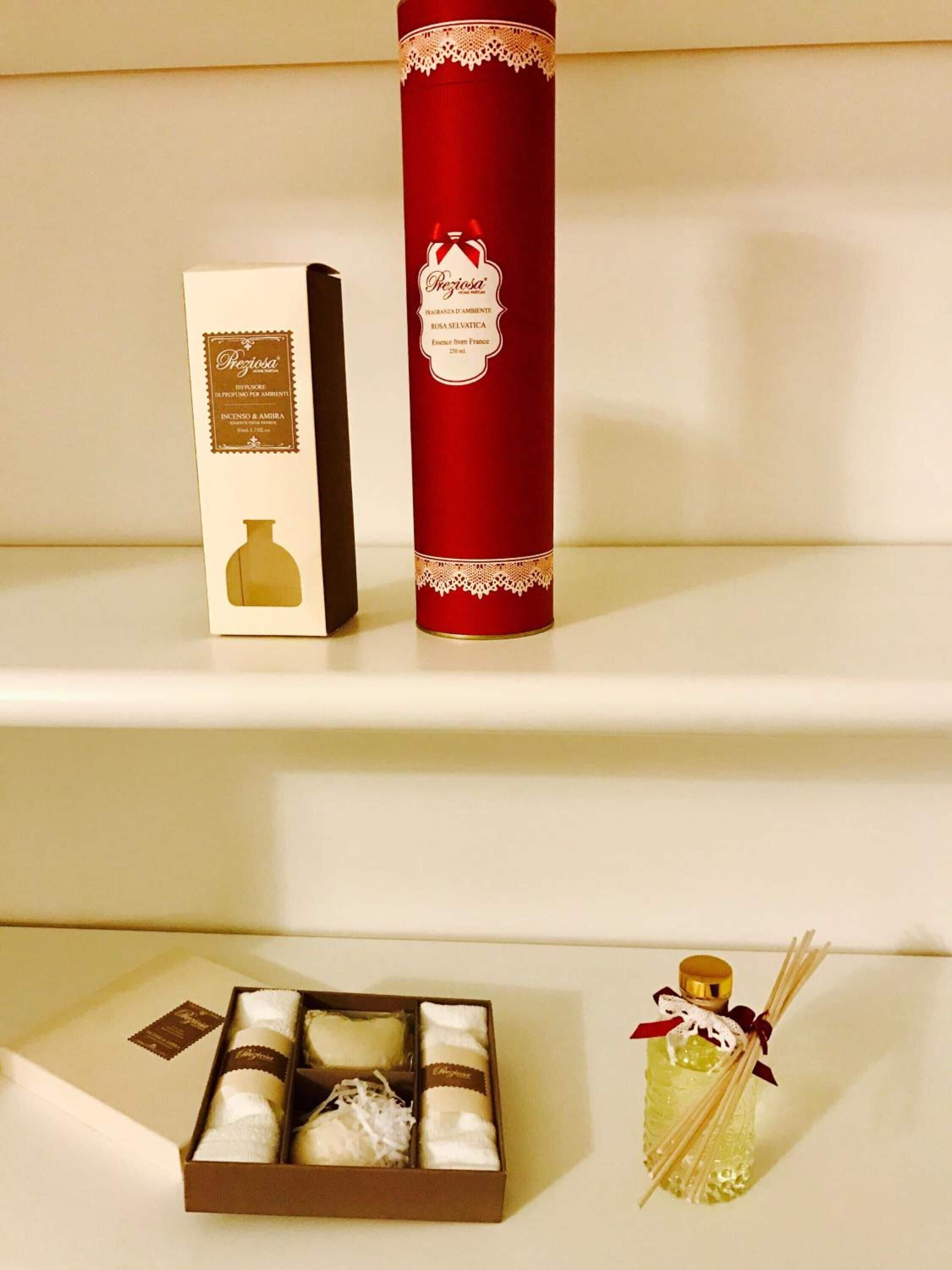 A shelf with a red cylindrical container labeled 'Preziosa' and a box with similar branding. Below, a box of scented candles and a bottle of reed diffuser are displayed.