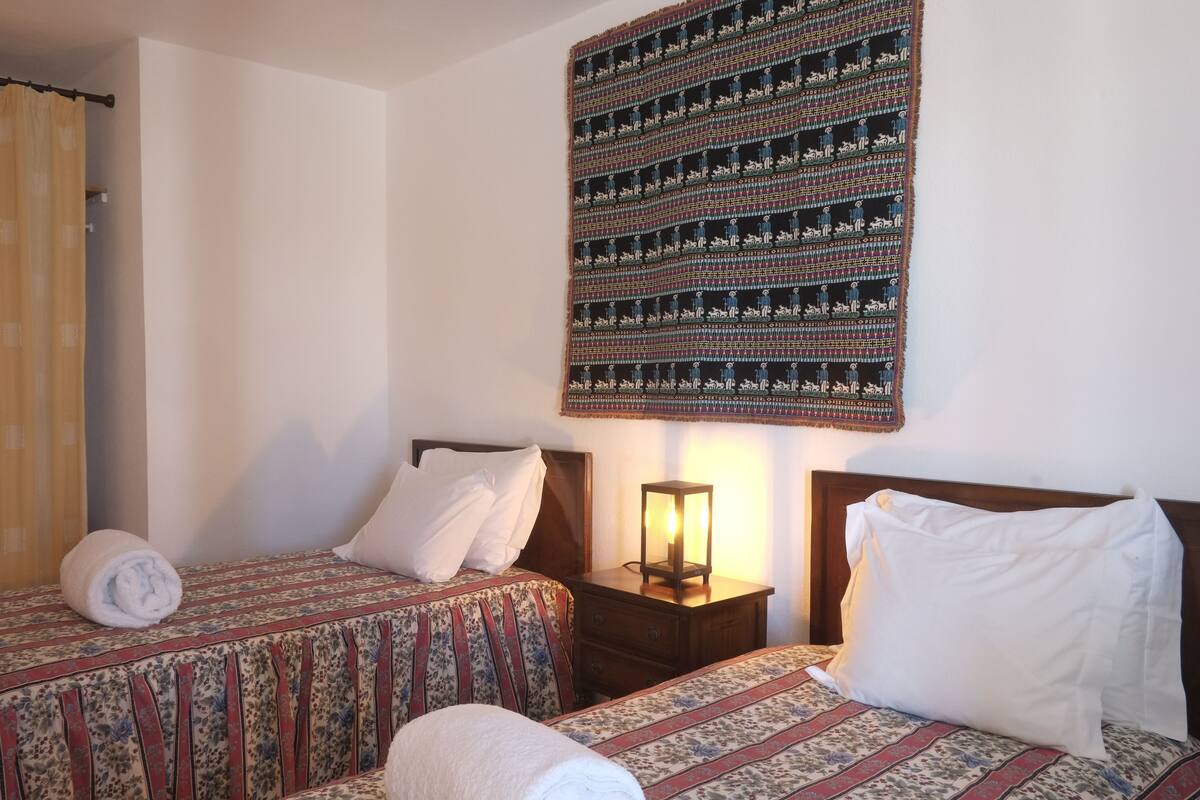 Ein Hotelzimmer mit zwei Betten, jeweils mit einem gemusterten Überwurf, einem Nachttisch mit einer Lampe und einem großen Wandteppich.