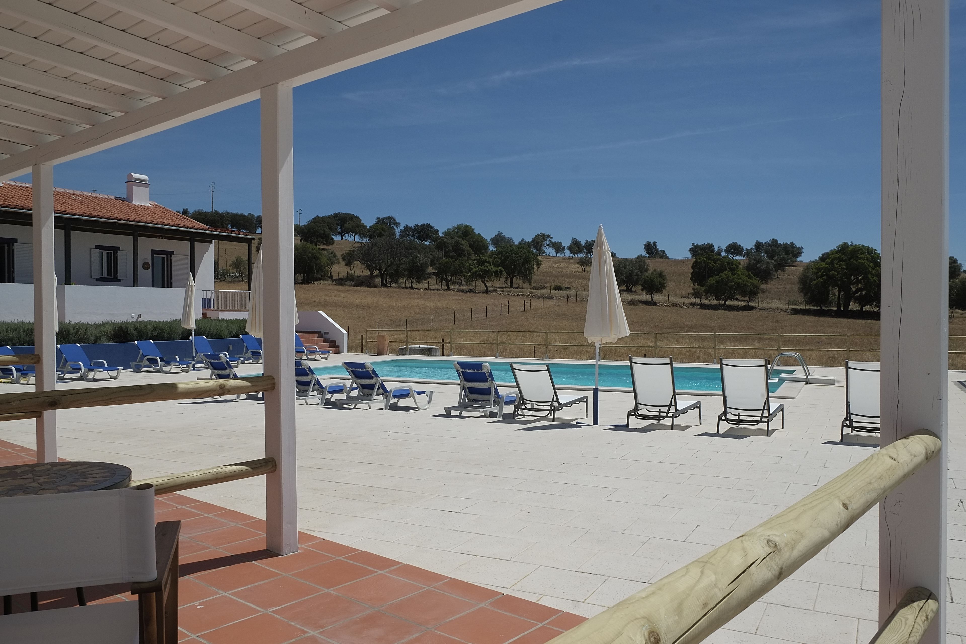 A poolside area with lounge chairs and umbrellas, a covered patio, and a view of a hilly landscape.