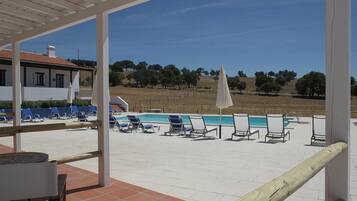 A poolside area with lounge chairs and umbrellas, a covered patio, and a view of a hilly landscape.