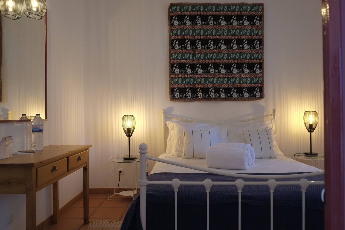 Ein ordentlich eingerichtetes Schlafzimmer mit Bett, Nachttischlampen, einem Holznachttisch und einem an der Wand hängenden Kunstwerk.
