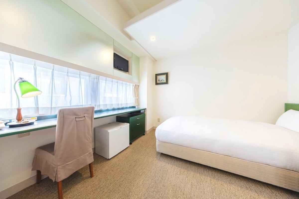 Habitación de hotel con cama, escritorio con silla, refrigerador pequeño, ventana con cortinas y televisión montada en la pared.