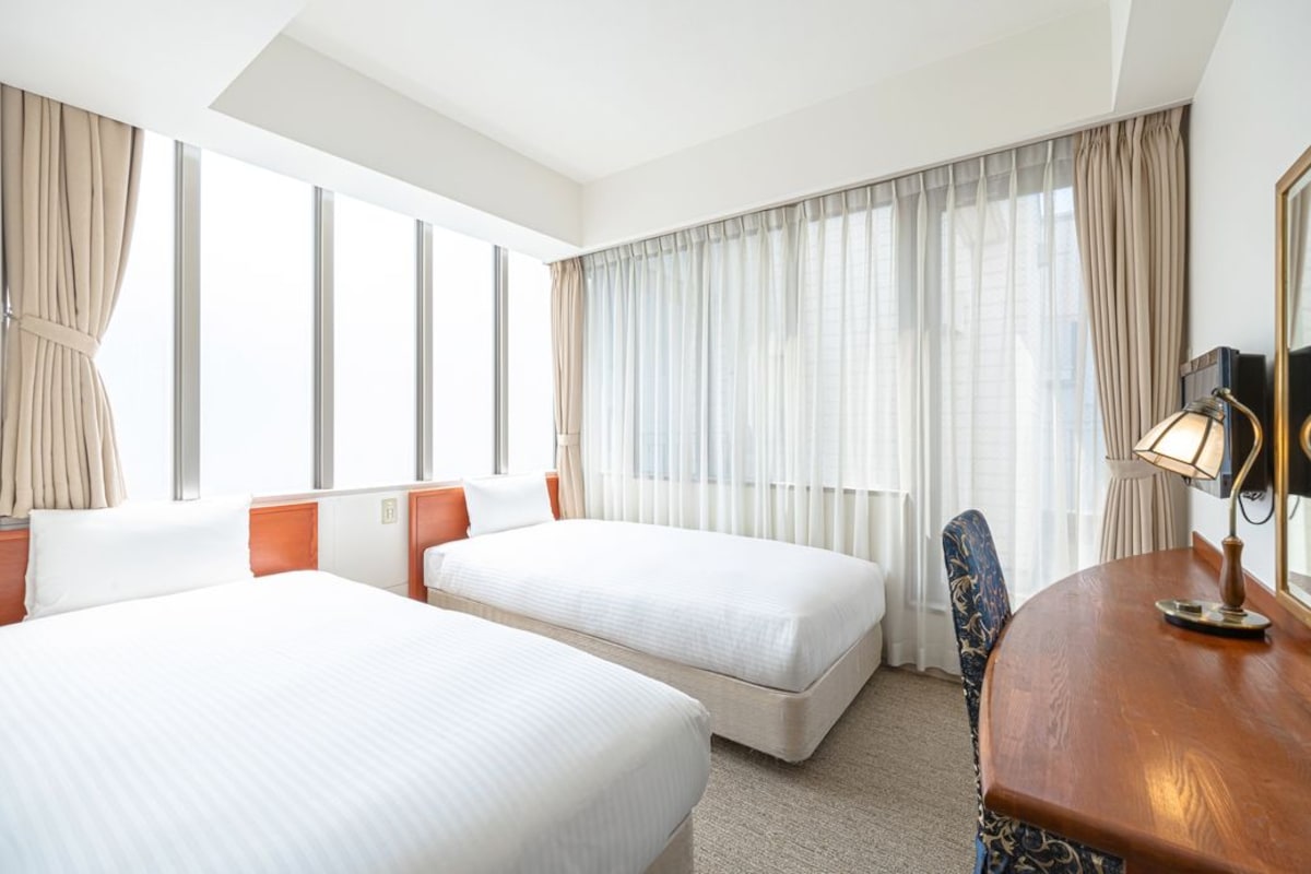 Habitación de hotel con dos camas, un escritorio, una silla y un amplio ventanal con cortinas.
