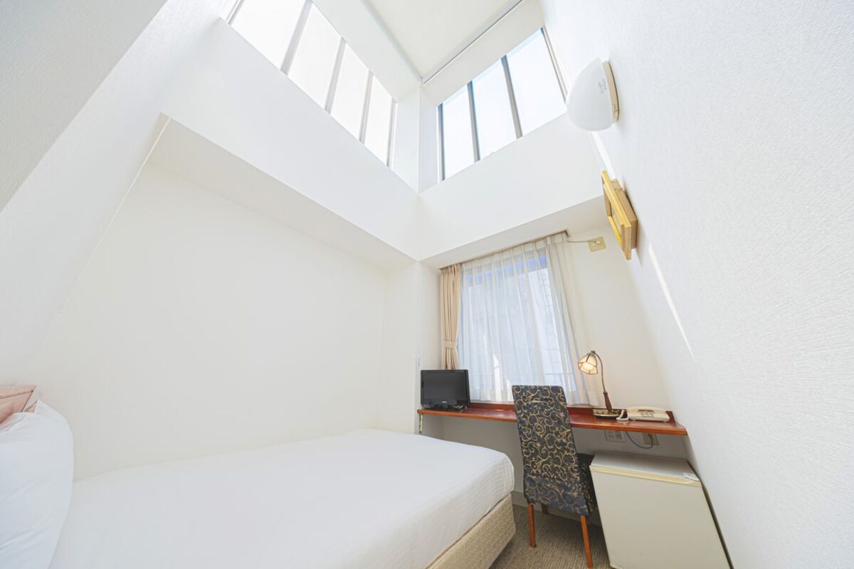 Habitación de hotel con cama, escritorio con computadora, silla, refrigerador pequeño y ventana con cortinas.