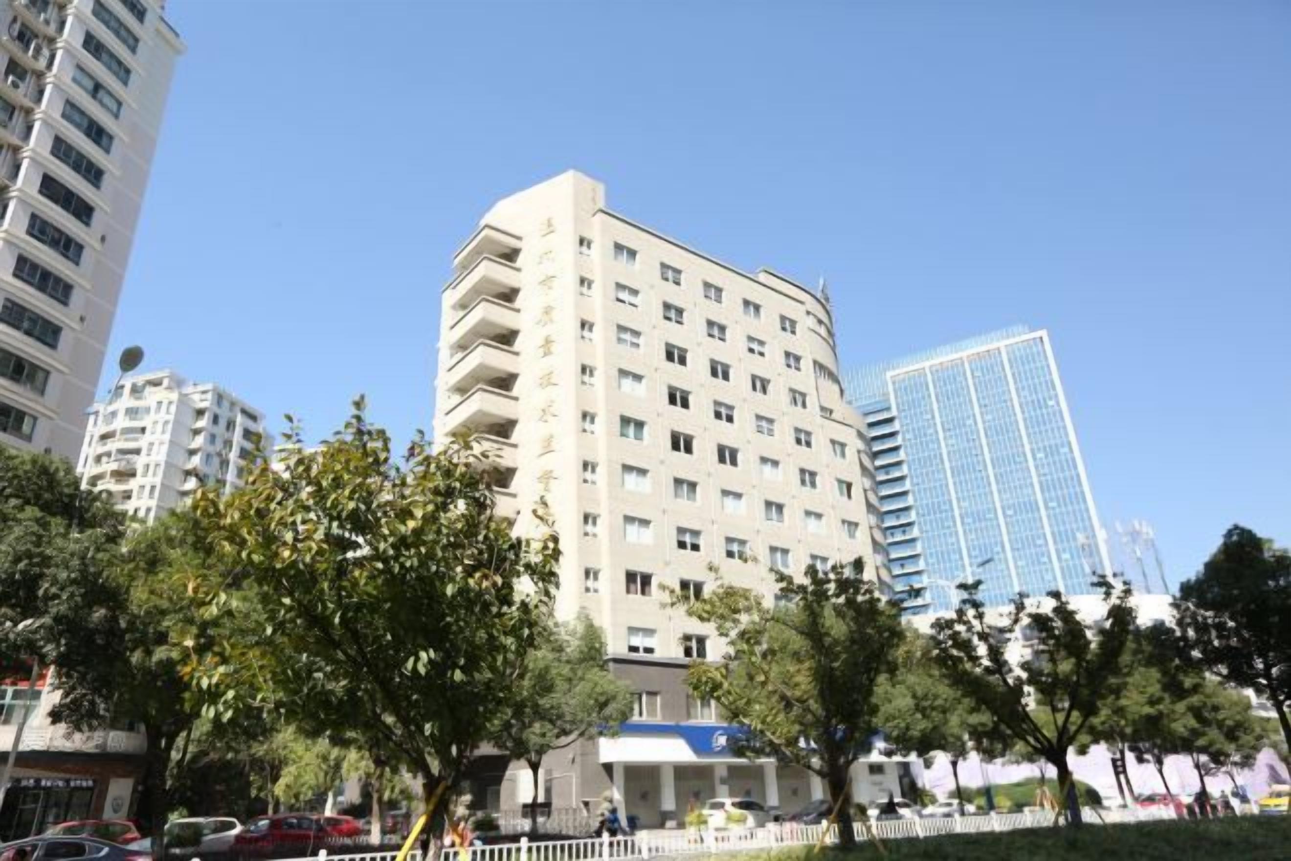 Un edificio de varios pisos con fachada plana y numerosas ventanas, rodeado de árboles y otros edificios.