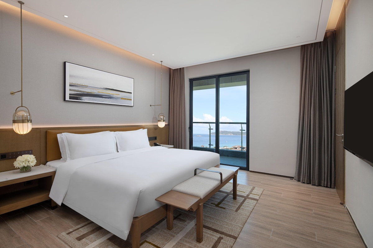 A modern hotel room with a large bed, bedside tables, a bench, a view of the sea, and a mounted TV.