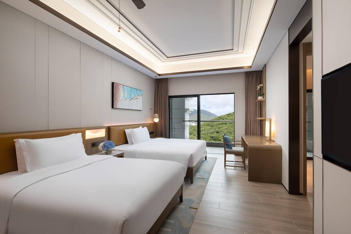 A hotel room with two beds, a desk, a TV, and a large window with a view of greenery.