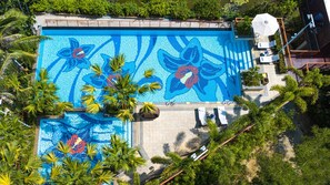 Vue aérienne d’une piscine ornée de mosaïques, entourée de palmiers et bordée d’une terrasse avec des chaises longues.