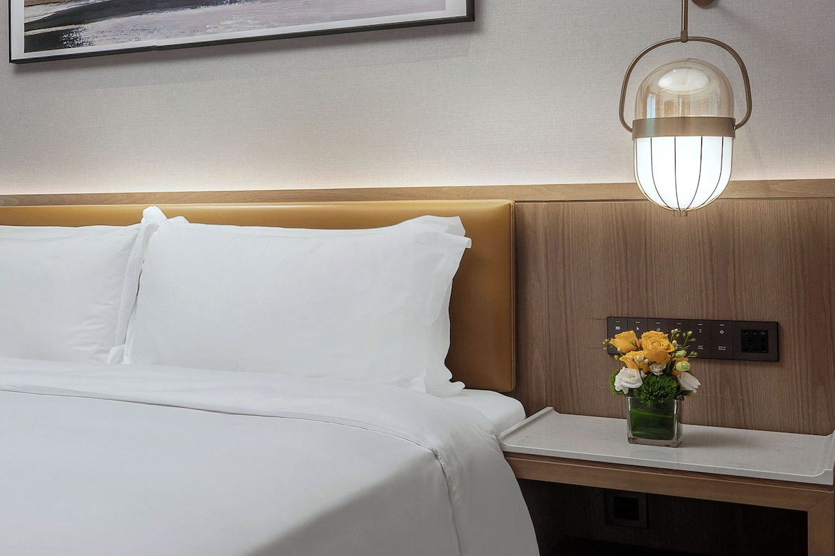 Una habitación de hotel con una cama, una mesita de noche con un jarrón de flores, una lámpara de pared y un enchufe.