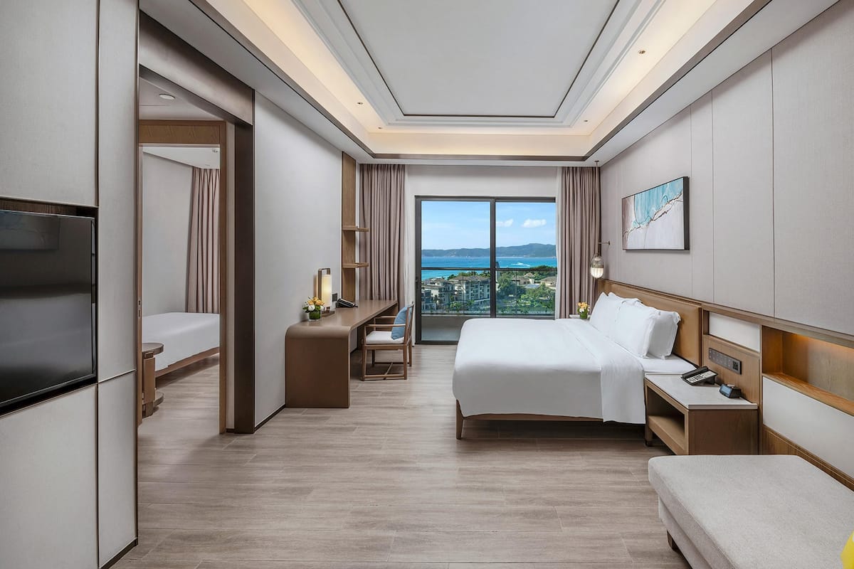 A hotel room with a bed, bedside tables, a TV, a desk, and a view of the city.