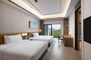 A hotel room with two beds, a desk, a TV, and a large window with a view of greenery.