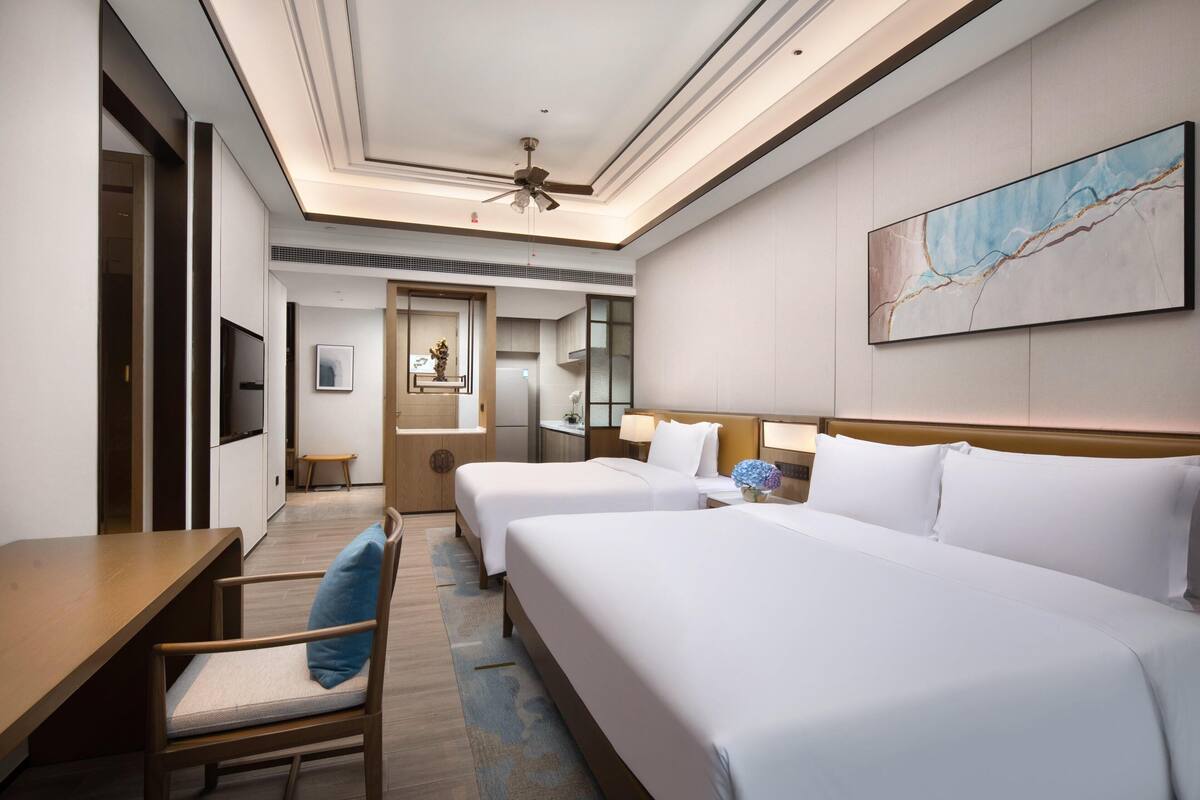 A hotel room with two beds, a desk, a chair, a TV, a ceiling fan, and a painting on the wall.