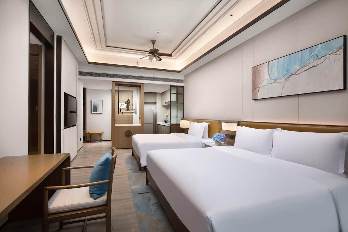 Quarto de hotel com duas camas, uma escrivaninha, uma cadeira, uma televisão, um ventilador de teto e um quadro na parede.