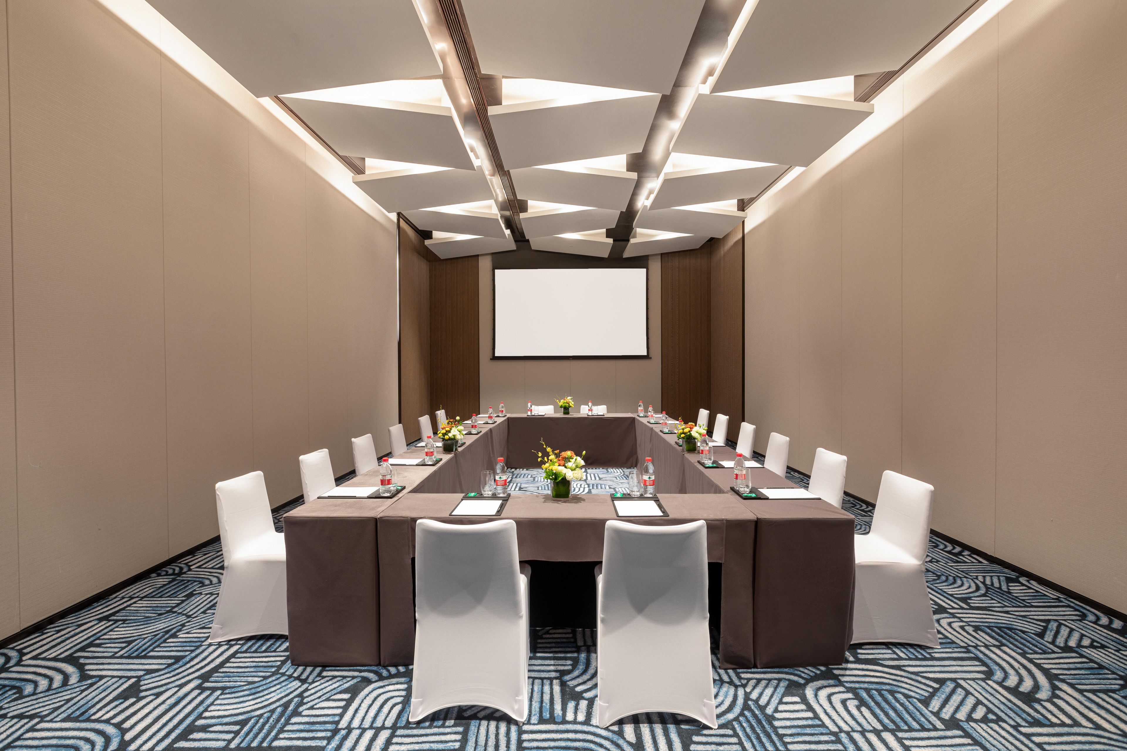 A conference room with a long table, white chairs, a projector screen, and a patterned carpet.