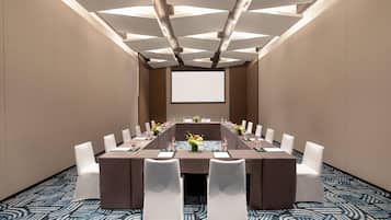 A conference room with a long table, white chairs, a projector screen, and a patterned carpet.