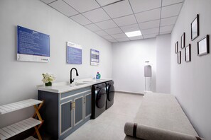 Un baño moderno con lavamanos, espejo y lavadora integrada. Se observan cuadros enmarcados en la pared y un pequeño estante con flores.