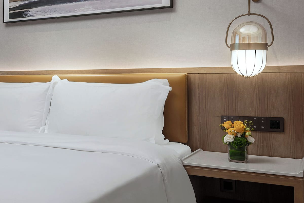 A hotel room with a bed, a nightstand with a vase of flowers, a wall-mounted light fixture, and a power outlet.