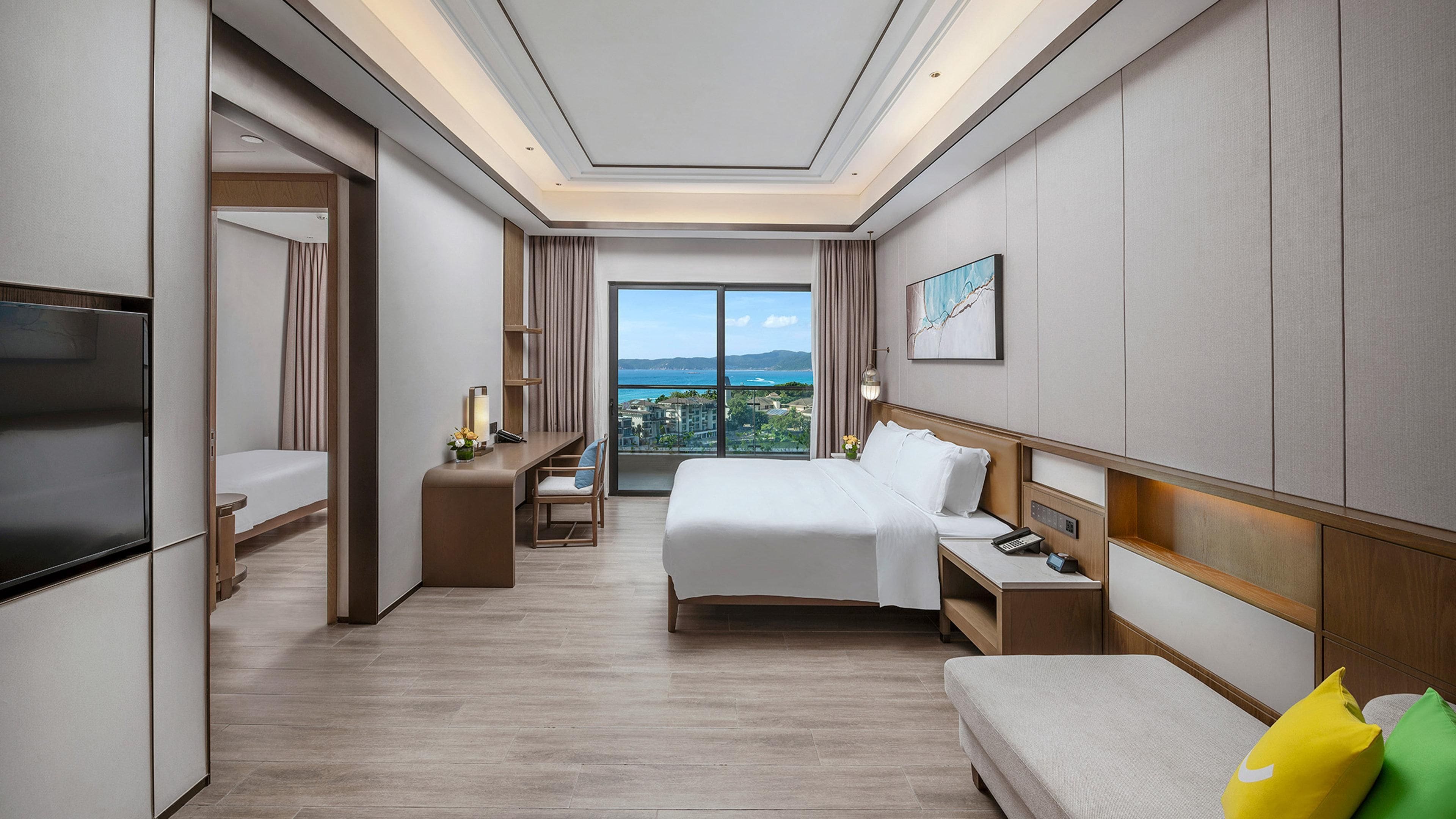 A hotel room with a bed, bedside tables, a TV, a desk, and a view of the city.