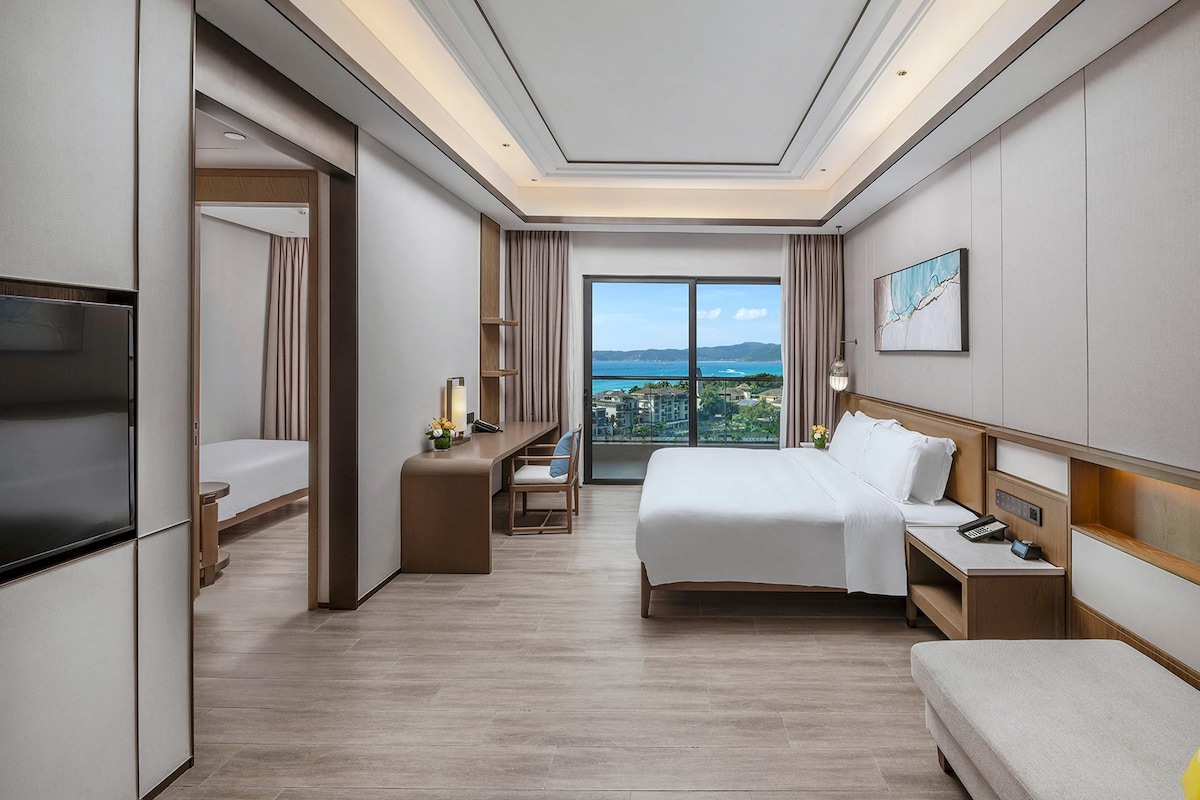 A hotel room with a bed, bedside tables, a TV, a desk, and a view of the city.